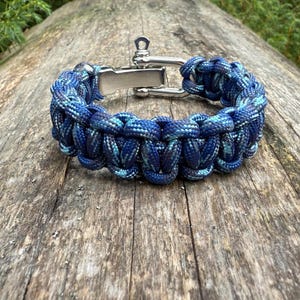 May include: A blue and teal paracord bracelet with a stainless steel clasp. The bracelet is made of tightly woven cord and is displayed on a wooden surface. The clasp is a silver color.