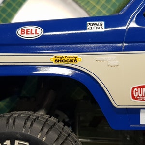 Pode incluir: Um caminhão modelo azul e branco com vários adesivos, incluindo "Bell", "Power Gloss", "Rough Country Shocks", "Custom", "Gumout" e "Direc".