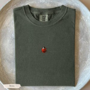 May include: A moss green t-shirt with a small embroidered ladybug. The shirt is folded neatly and has a crew neck. The tag reads "Comfort Colors". The shirt is displayed on a light gray, textured surface.