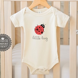 May include: Cream baby bodysuit featuring a red and black ladybird design and the words "little bug" in black script. The garment hangs on a wooden hanger in front of a white cot.