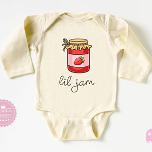 Pode incluir: Um body de bebê creme com mangas compridas. A frente apresenta uma ilustração de desenho animado de um pote de geleia de morango com um morango no rótulo. As palavras "lil jam" estão escritas abaixo do pote em itálico.