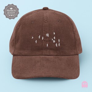 May include: A brown corduroy baseball cap with a curved brim. The cap features a white embroidered design of birds in flight. A "Premium Quality" badge is visible on the left side. The cap is set against a light blue background.