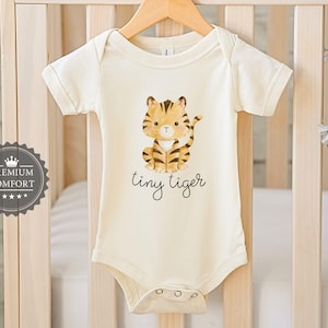 May include: Cream-coloured baby bodysuit featuring a watercolour illustration of a tiger cub and the words "tiny tiger" in script. The bodysuit is hanging on a wooden hanger in front of a cot.