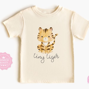 May include: A cream-coloured t-shirt with a watercolour illustration of a cartoon tiger with brown stripes. The text "tiny tiger" is written in a cursive font below the tiger. The shirt has short sleeves and a crew neck.