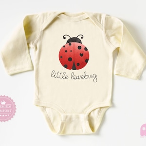 May include: Cream-colored baby bodysuit with long sleeves. The front features a red ladybug design with black spots and the words "little lovebug" in a handwritten font. A pink seal with the words "Premium Comfort" is on the left.