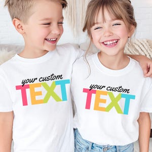 May include: Two children wearing white t-shirts with the text "your custom TEXT" in a colorful, blocky design. The text is in black, pink, orange, green, and blue. The children are smiling and appear happy.