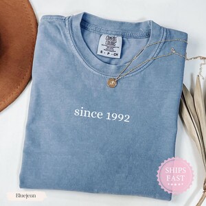 May include: Bluejean-colored Comfort Colors t-shirt with the text "since 1992" in white. A gold necklace with a pendant rests on the shirt. The shirt is folded and has a tag at the neckline.