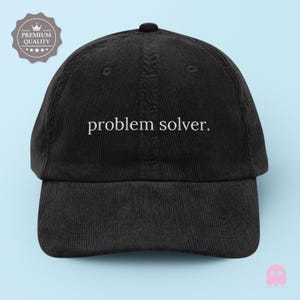 May include: Black corduroy baseball cap featuring the embroidered phrase "problem solver." The cap has a curved peak and a button on top. A "Premium Quality" emblem is visible on the left side.