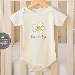 May include: Cream baby bodysuit featuring a daisy graphic and the text "lil daisy". The daisy has white petals and a yellow, smiling centre. The bodysuit is hanging on a wooden hanger in front of a cot.