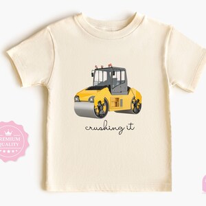 Op de afbeelding: Crèmekleurig T-shirt met een gele wals met zwarte wielen en een grijze cabine. De tekst "crushing it" staat in cursiefschrift eronder. Een leuk, speels ontwerp voor kinderen.