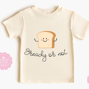 May include: Cream-colored t-shirt featuring a smiling cartoon bread slice with arms. The text "Bready or not" is written in a whimsical script below the bread illustration. A pink premium quality badge is on the left.