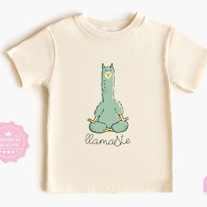 Op de afbeelding: Crèmekleurig T-shirt met een cartoon lama in een yogahouding, in groen- en geeltinten. Het woord "llamaste" is geschreven in een handschrift onder de lama. Het shirt heeft korte mouwen en een ronde hals.