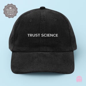 May include: Black corduroy baseball cap with the embroidered text "TRUST SCIENCE" in white. The cap has a curved brim and a button on top. A "Premium Quality" badge is in the upper left corner.