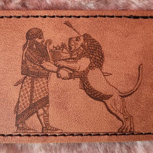 Puede incluir: Un parche rectangular de cuero marrón con un borde cosido en negro. El parche presenta un grabado detallado de una figura frente a un león. La figura sostiene la melena del león, y el león se alza sobre sus patas traseras.