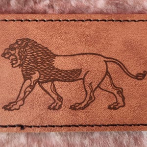 Puede incluir: Un parche rectangular de cuero marrón claro con una ilustración marrón de un león. El león se representa de perfil, caminando con la cabeza girada. El parche tiene un borde cosido negro.