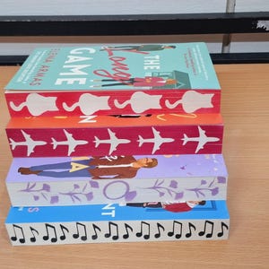 May include: A stack of five paperback books with colorful covers. The top book has a light blue cover with the text "The Long Game" in red and white. The second book has a red cover with the text "In" in white. The third book has a pink cover with a pattern of airplanes. The fourth book has a purple cover with a floral pattern. The bottom book has a light blue cover with a pattern of musical notes.