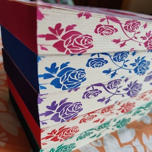 May include: A stack of five books with white covers and a repeating pattern of pink, blue, purple, and red roses. The books are stacked on a patterned surface.