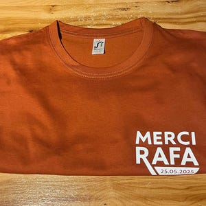 Peut inclure: T-shirt de couleur orange brûlé avec l'inscription blanche "MERCI RAFA" et la date "25.05.2025". Le t-shirt a un col rond et une petite étiquette avec un logo.
