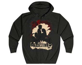 May include: Black zip-up hoodie featuring a silhouette design with the word "HADESTOWN" in a distressed font. The graphic includes figures and a red and beige background.
