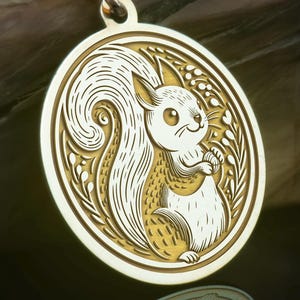 Könnte beinhalten: Goldfarbene Halskette mit einem Anhänger, der ein Eichhörnchen-Design in einem Kreis zeigt. Das Eichhörnchen ist in Weiß und Gold gehalten und hält eine Nuss. Der Anhänger hängt an einer feinen Goldkette. Ein Schmuckstück mit Naturmotiv und aufwendigen Details.