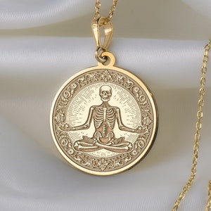 Op de afbeelding: Een goudkleurige hangerketting met een gedetailleerde illustratie van een skelet in een zittende yogahouding. De ronde hanger heeft een sierlijke rand en hangt aan een delicate ketting. De achtergrond is een zachte, witte stof.
