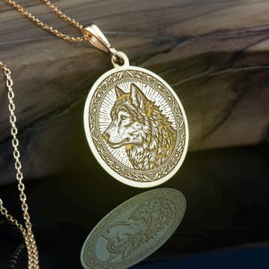 Può includere: Una collana con un ciondolo in oro che presenta un'immagine incisa dettagliata di una testa di lupo. Il lupo è rivolto a destra e ha un motivo di pelliccia dettagliato. Il ciondolo è rotondo e ha un bordo decorativo.