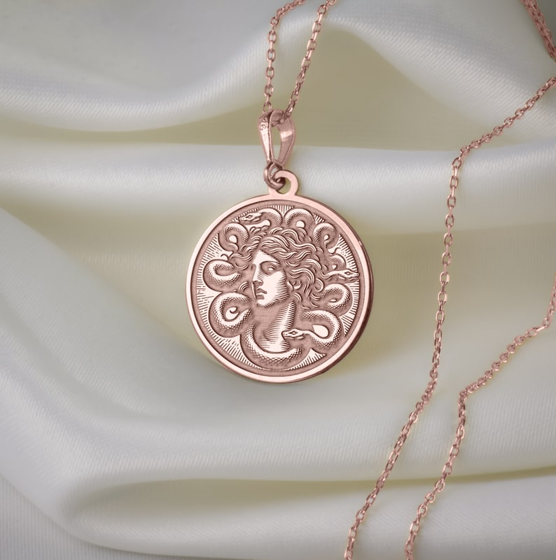 May include: A rose gold necklace with a round pendant featuring a detailed engraving of Medusa's head surrounded by snakes.