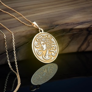 May include: A gold necklace with a round pendant featuring a detailed engraving of Medusa's head surrounded by snakes. The pendant is hanging on a chain against a wooden background.