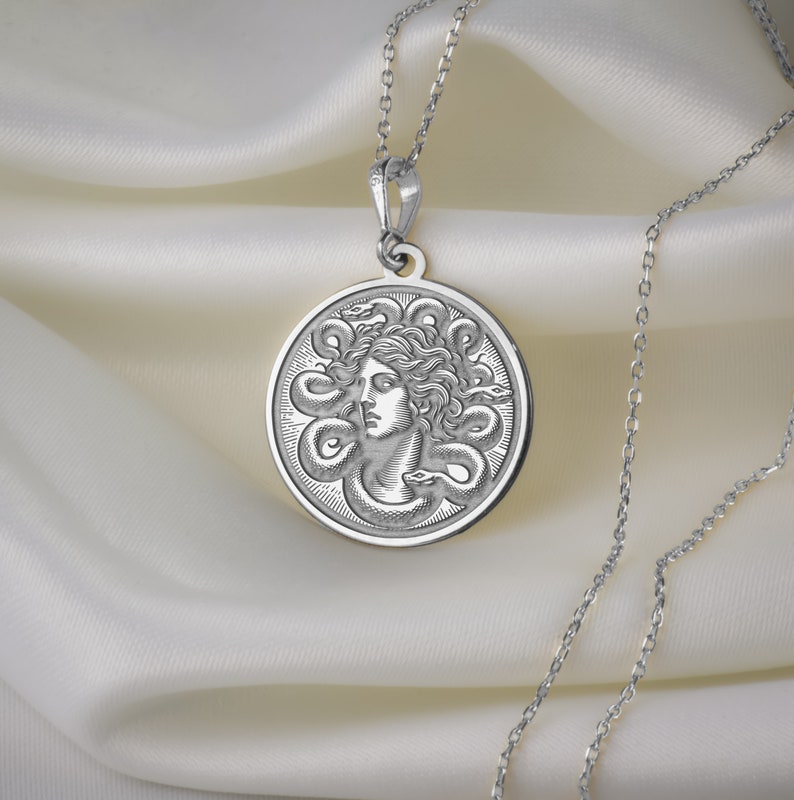 May include: Silver necklace with a round pendant featuring a detailed engraving of Medusa's head surrounded by snakes.