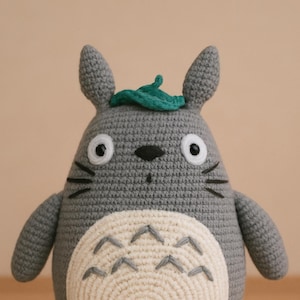 Puede incluir: Peluche de Totoro tejido a ganchillo, gris con vientre crema. El peluche tiene ojos negros, bigotes y una hoja verde en la cabeza. El texto "CROCHET PATTERN" está en la parte inferior de la imagen.