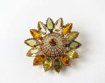 Peut inclure: Une broche circulaire dotée d'un dôme central en métal doré en forme de cage avec une pierre ambrée au sommet. Le dôme est entouré d'un motif rayonnant de pierres facettées taille marquise dans des tons de vert olive et d'ambre.