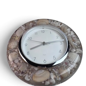 May include: A round wall clock featuring a white face with black numerals and silver hands. The frame is made of clear resin embedded with a variety of natural seashells in shades of beige, tan, and white. The brand name Krich is printed in black.