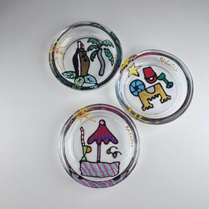 May include: Three clear glass dishes featuring hand-painted designs. One dish shows a palm tree and a bottle, another displays a sandcastle with a bucket and shovel, and the third depicts a beach umbrella with a drink and sunglasses. Each is signed RAENGLL.