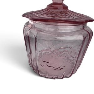 May include: A translucent purple glass jar with a matching lid. The jar features a faceted, hexagonal body with embossed floral patterns on the sides. The lid has a scalloped edge and a central knob, also decorated with intricate floral relief details.