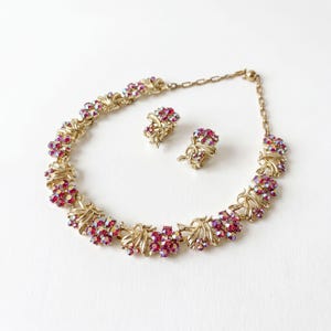 May include: A gold-tone metal necklace and matching clip-on earrings set. The jewelry features clusters of iridescent pink and red faceted crystals arranged in floral-inspired shapes, connected by gold-tone metal links and a chain.