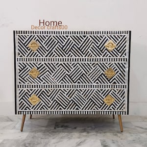 May include: A three-drawer dresser with a black and white geometric pattern made of mother of pearl inlay. The dresser has gold-toned hardware and tapered legs.