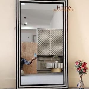 Puede incluir: Un espejo grande y rectangular con un marco negro y un borde de incrustaciones de hueso blanco. El espejo refleja una habitación con un armario de madera y una mesa con una tapa de incrustaciones de hueso.