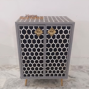 May include: A gray cabinet with a geometric honeycomb pattern on the doors and top. The cabinet has brass-colored handles and light-colored wooden legs. The cabinet is a decorative piece of furniture.