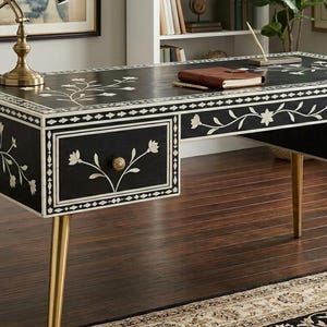以下が含まれることがあります： 白のフローラルインレイと金色の脚が付いた黒いデスク。デスクには2つの引き出しと装飾的なフローラルパターンがあります。茶色のノートとペンがデスクの表面に置かれています。家具です。