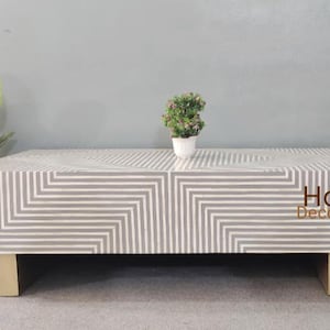 May include: A white and gray geometric patterned coffee table with gold metal legs. The table is made of bone inlay and has a modern design.
