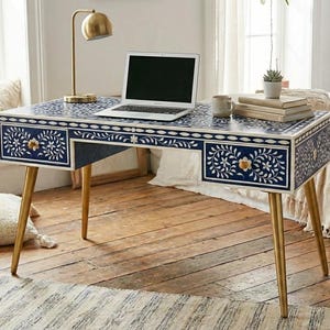 以下が含まれることがあります： 金色の脚が付いた、青と白の花柄のデスク。 デスクには金色のノブが付いた2つの引き出しがあり、上にはラップトップがあります。 金色のデスクランプとマグカップもデスクにあります。 デスクは窓のある明るい部屋にあります。