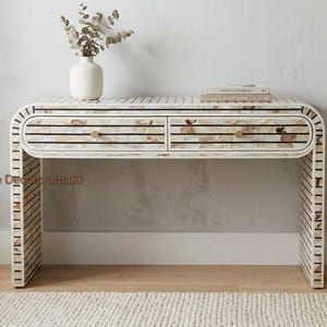 Peut inclure: Une console avec un cadre incurvé et deux tiroirs, présentant un motif incrusté de tuiles en coquillage irisé séparées par de fines lignes horizontales noires. Chaque tiroir possède un petit bouton rond en métal doré. Le texte Home Decorcrafts00 est visible.