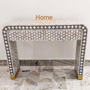 May include: A decorative console table with a geometric pattern. The table has two drawers and is adorned with a mosaic-like design in white, grey, and gold. The legs have gold accents at the base.