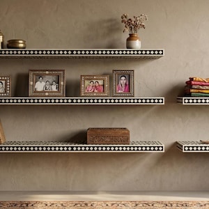 Può includere: Cinque mensole a muro sospese caratterizzate da un motivo geometrico a rombi bianco e nero lungo il bordo anteriore. Le mensole sono disposte in modo sfalsato e ospitano vari oggetti decorativi, tra cui vasi in metallo, foto incorniciate, libri e tessuti.