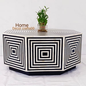 Op de afbeelding: Achtekantige salontafel met een zwart-wit geometrisch patroon gemaakt van beeninleg. De tafel heeft een zwarte basis en staat op een wit oppervlak.