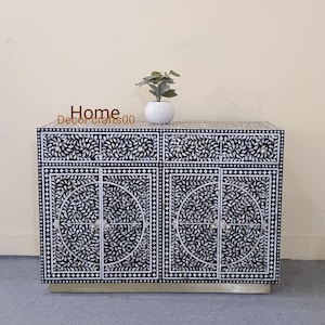 May include: Black and white cabinet with intricate floral patterns made from mother of pearl inlay. The cabinet has two drawers and two doors. The cabinet is sitting on a silver base.