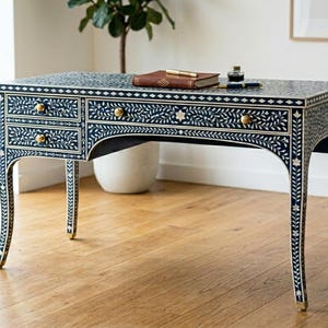 Può includere: Una scrivania blu scuro con intarsi floreali bianchi e ferramenta dorata. La scrivania ha tre cassetti e gambe curve. Un libro marrone, una penna e un calamaio sono sopra. La scrivania è larga circa 122 cm.
