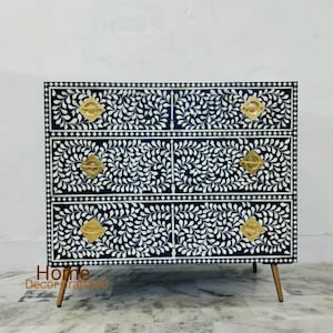 May include: A three-drawer dresser with a black and white bone inlay design. The drawers have gold-toned handles and the dresser is on four gold-toned legs.