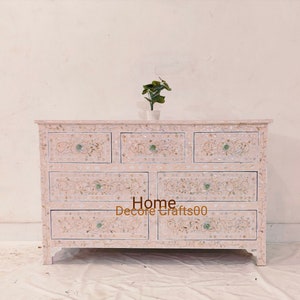 May include: A white dresser with six drawers, each decorated with intricate floral patterns and mother-of-pearl inlay. The dresser is made of wood and has a light pink finish. The dresser is sitting on a white floor. Home Decors Crafts00 is written on the front of the dresser.