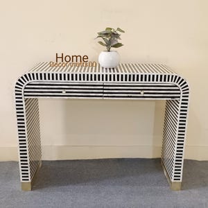 May include: A black and white striped console table with two drawers and gold-colored feet. The table has a curved design and a small white vase with a plant on top. The text "Home Decor crafts 500" is visible on the top.